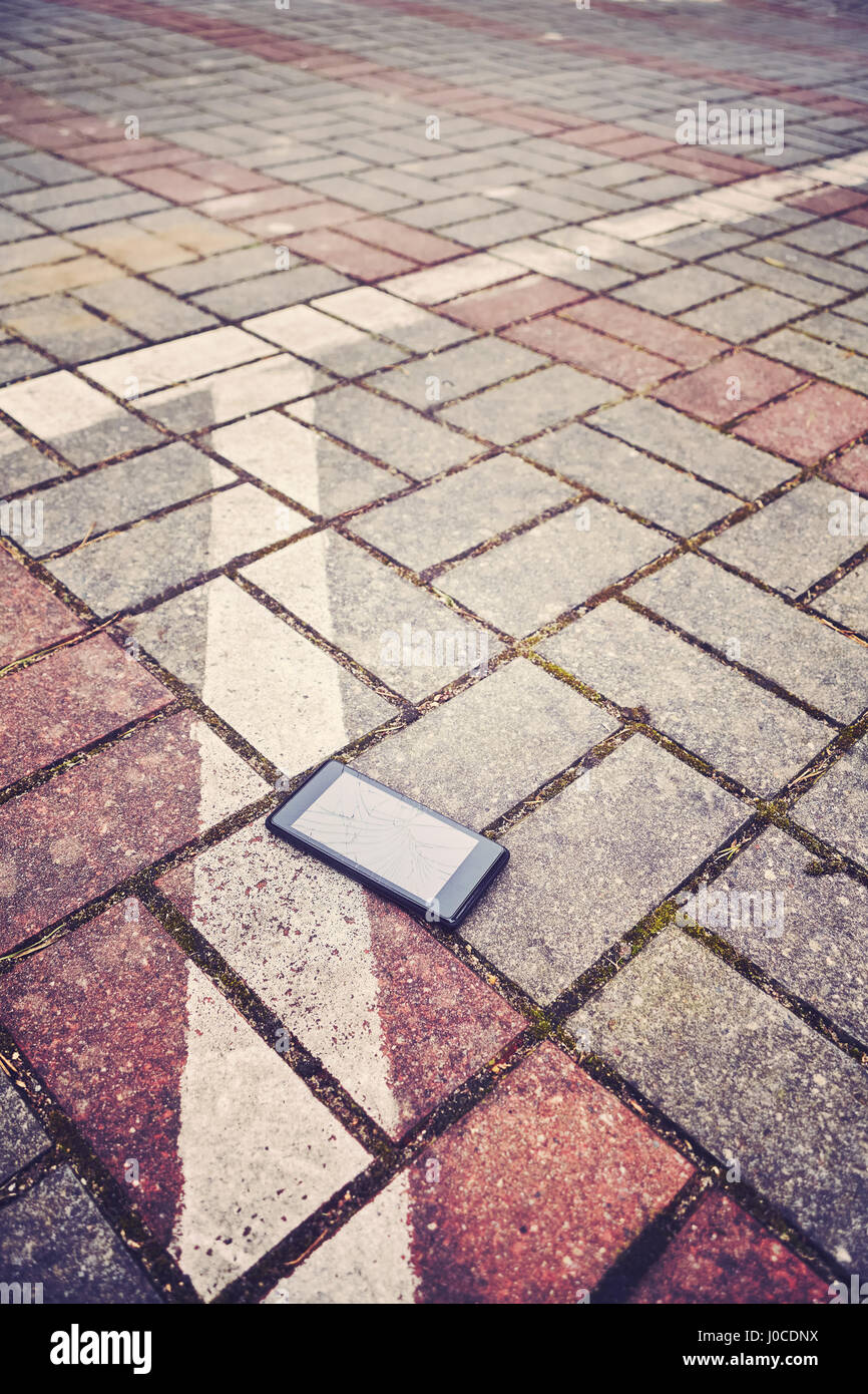 Vintage stylized picture of a mobile phone with broken screen on a parking pavement, selective focus. Stock Photo