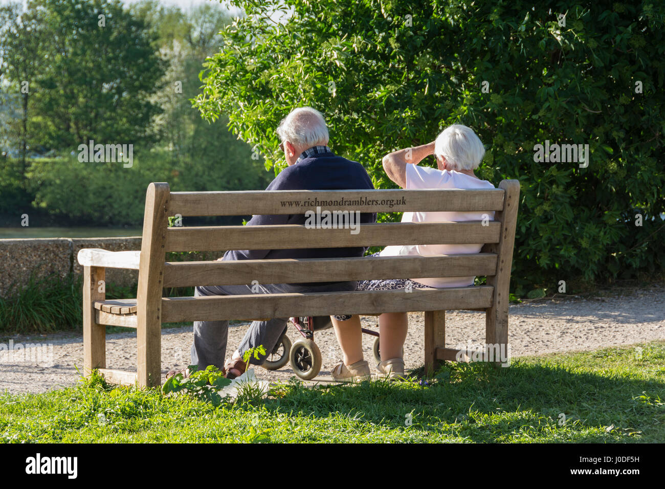 An elderly couple sitting on a park bench beside on the towpath beside the River Thames in London, England, UK Stock Photo