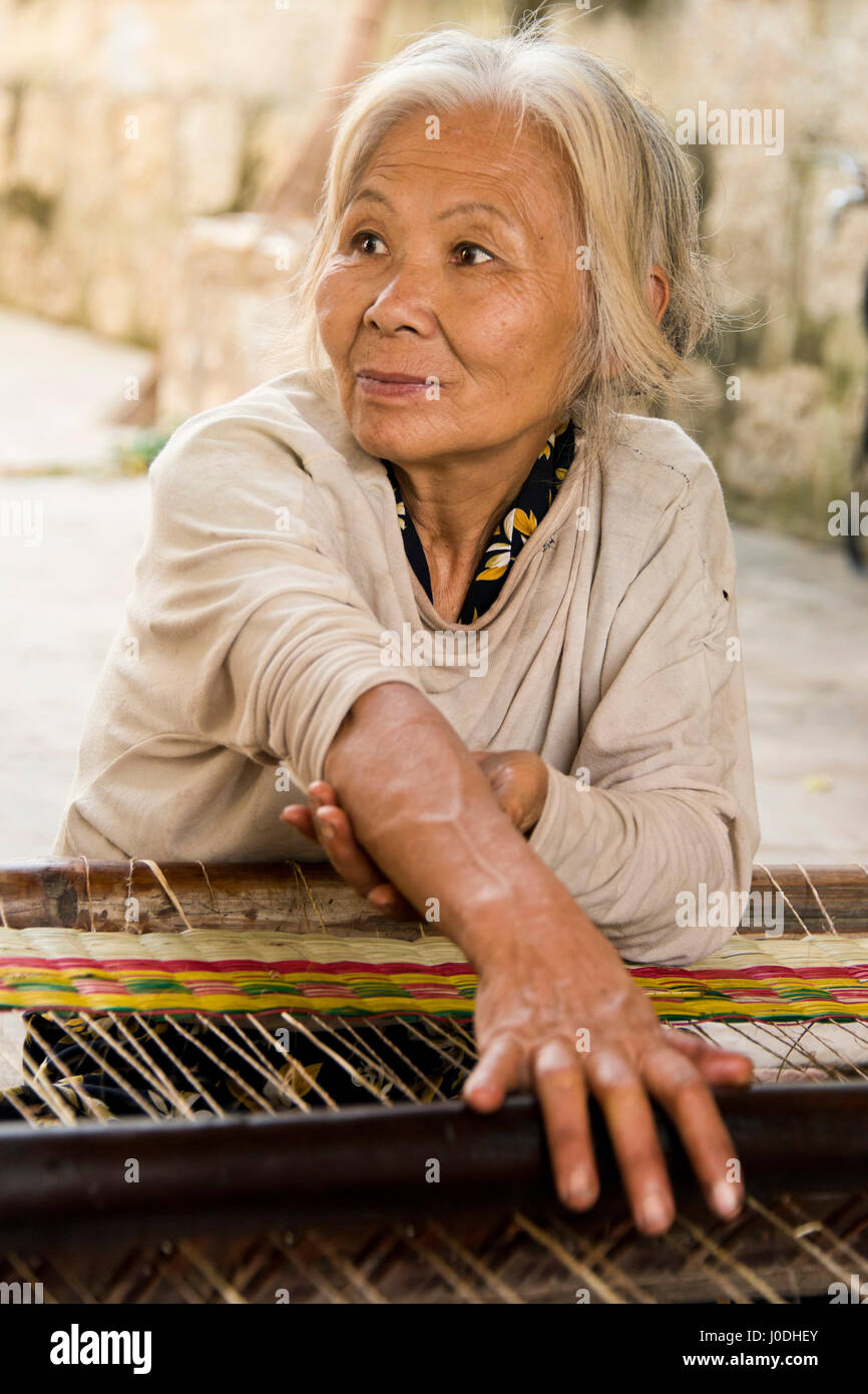 Vertical portrait of an old lady weaving traditional Vietnamese sleeping mats on a loom. Stock Photo