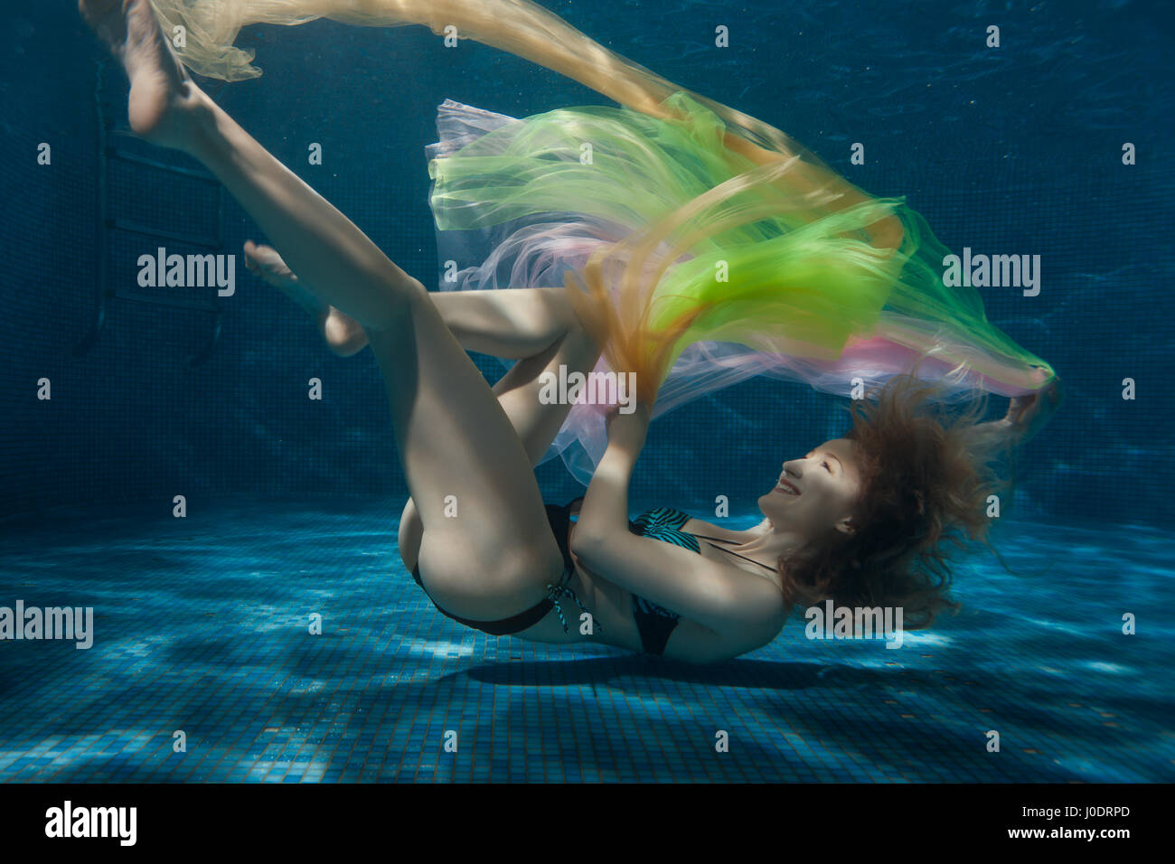 Woman lies on the bottom of the pool and playing with colorful fabrics. Stock Photo