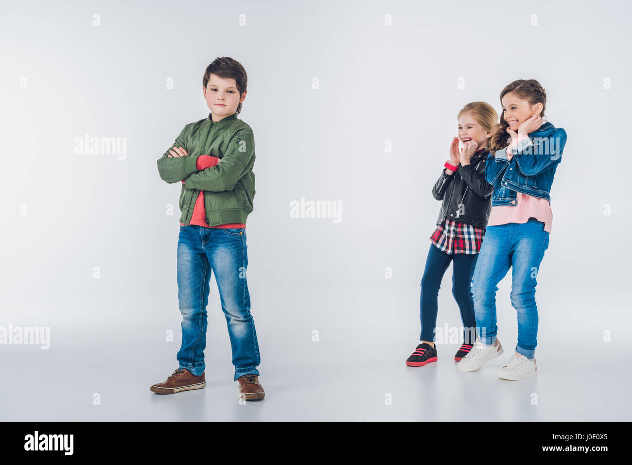 Two laughing girls making fun of boy standing with crossed arms on grey