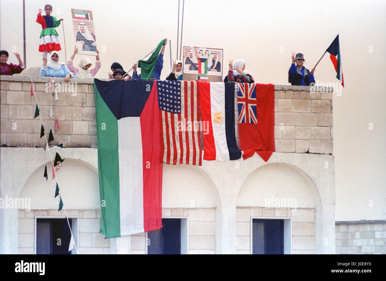 Kuwaitis wave flags and celebrate from the rooftops as coalition forces enter the city during the liberation of Kuwait from Iraq March 1, 1991 in Kuwait City, Kuwait. After four days of fighting, all Iraqi troops were expelled from Kuwait, ending a nearly seven-month occupation of Kuwait by Iraq. Stock Photo