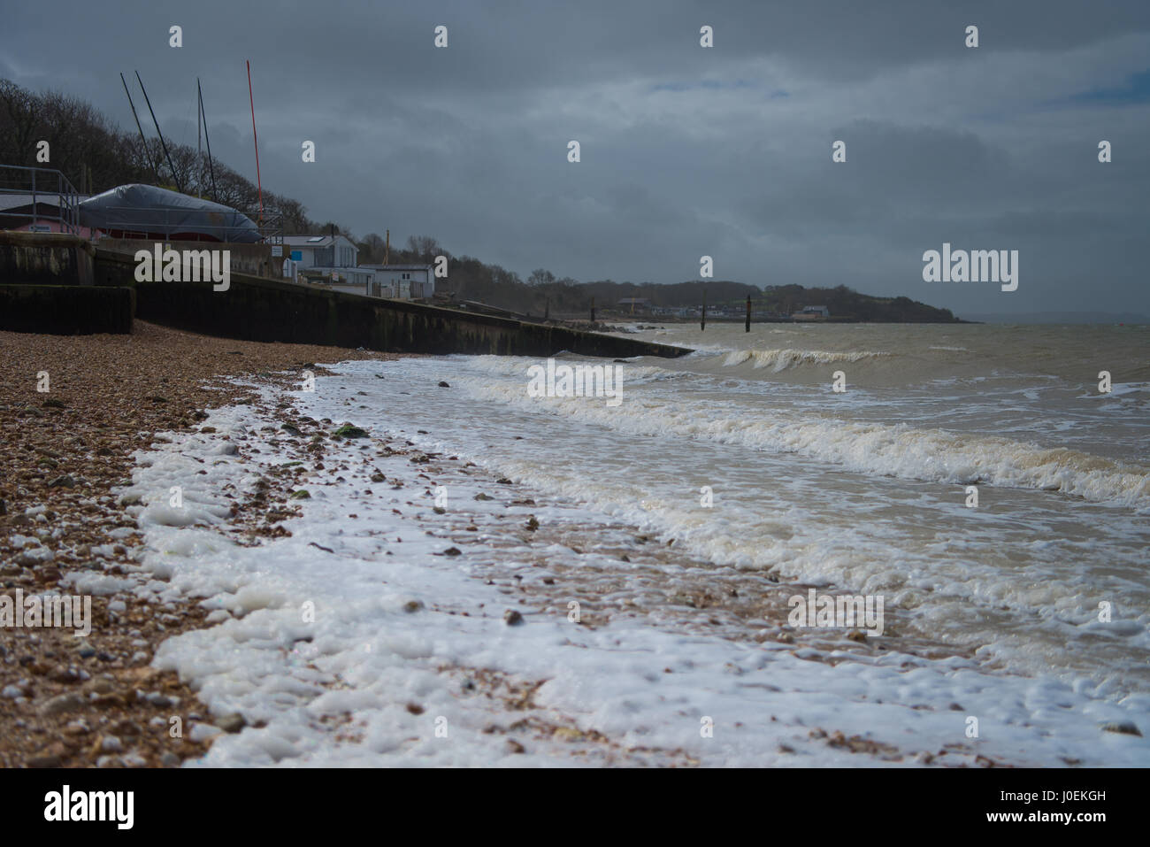 Stormy sea conditions with foam water and overcast sky, beach view with stones, a slipway, views of dinghy masts, distant buildings in bay. Stock Photo