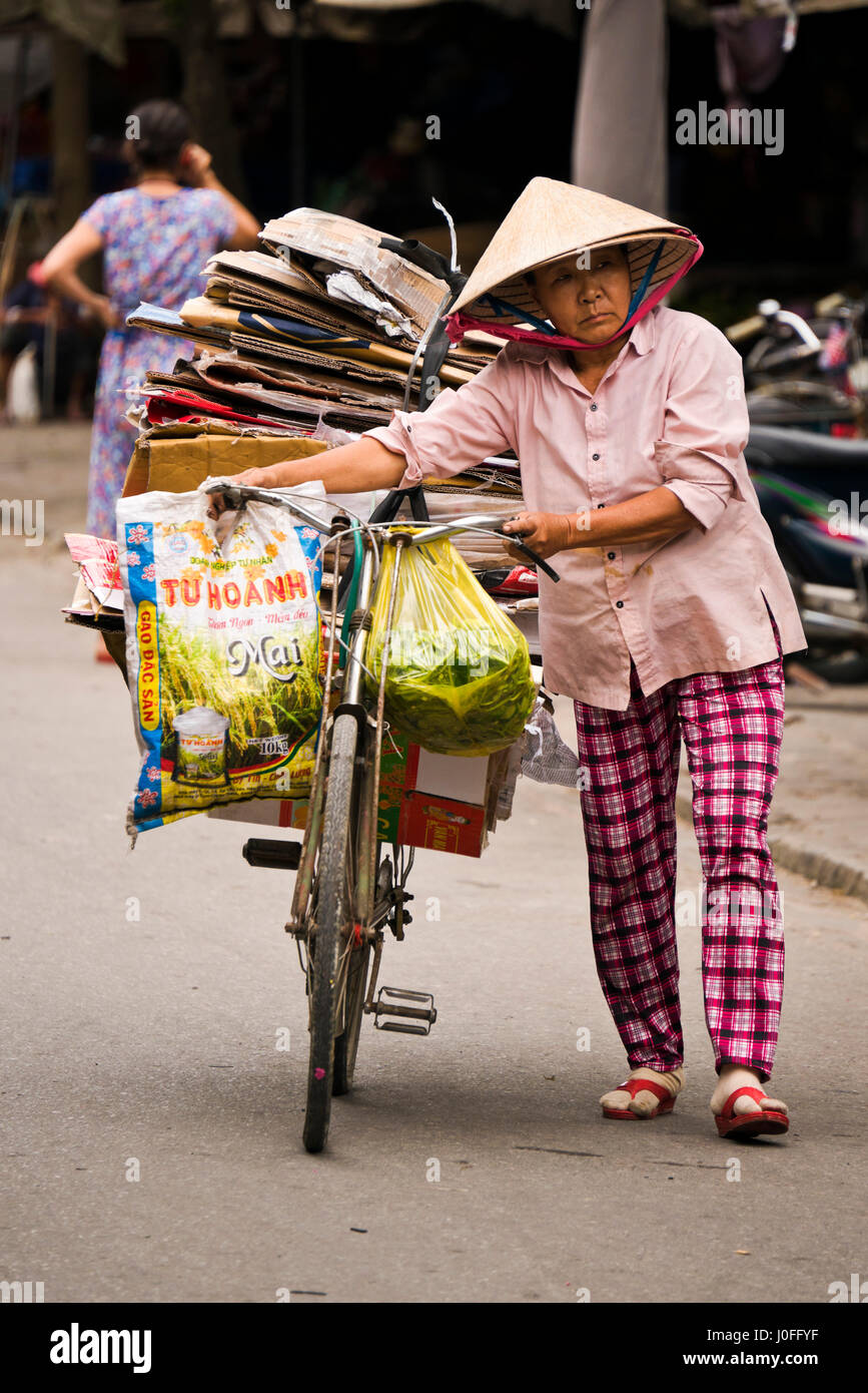 Vertical portrait of an old lady pushing a bicycle laden with cardboard boxes through the streets in Hoi An, Vietnam. Stock Photo