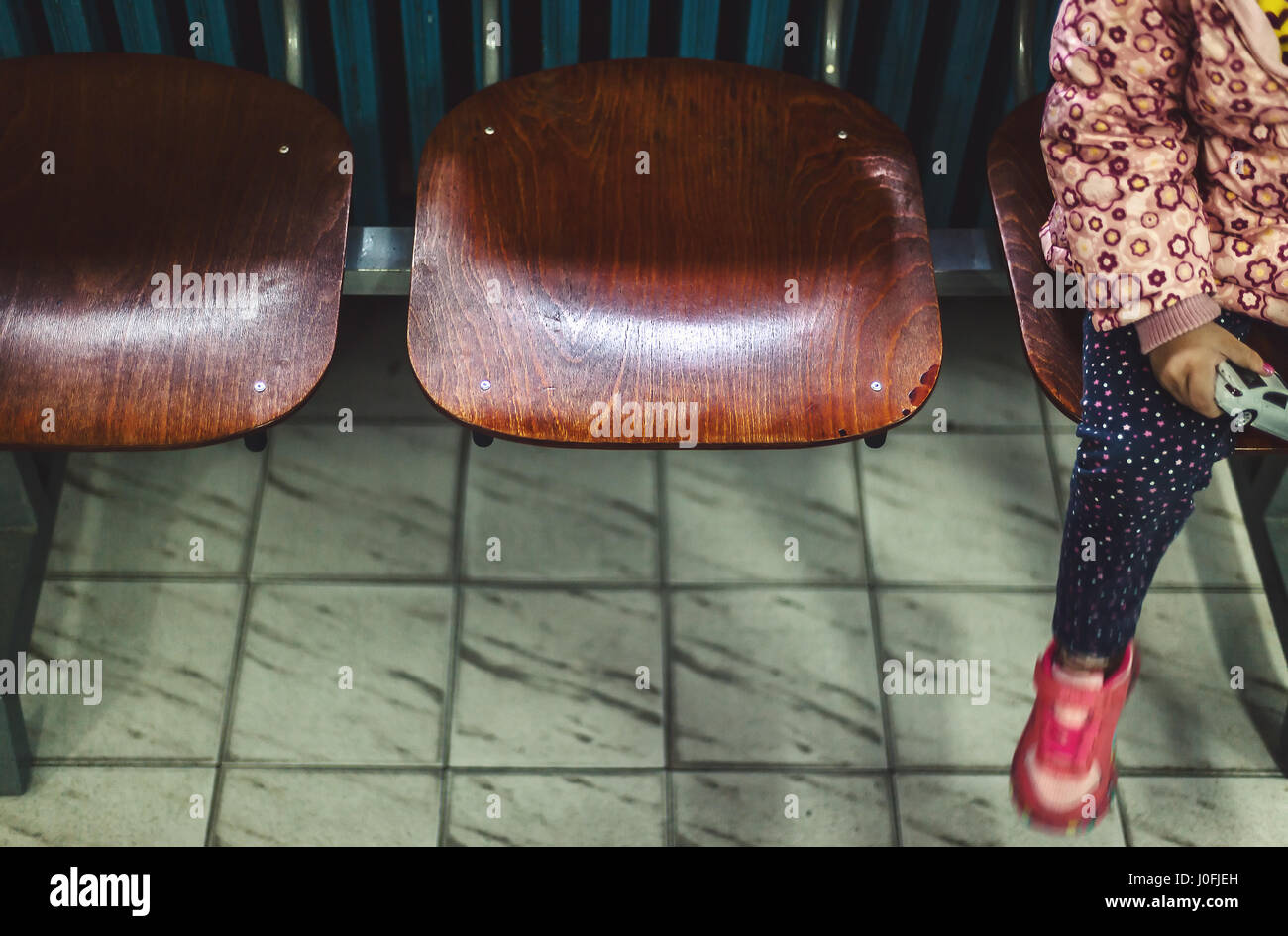 Three wooden chairs in waiting room and part of a body of a small child sitting and waiting. Stock Photo