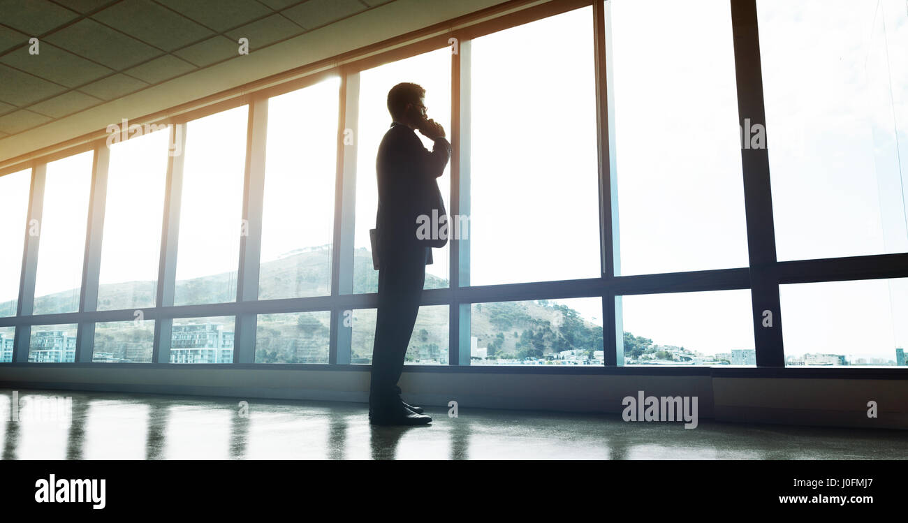 Full length of entrepreneur standing by window and talking on mobile phone. Man looking outside the office window and making a phone call. Stock Photo