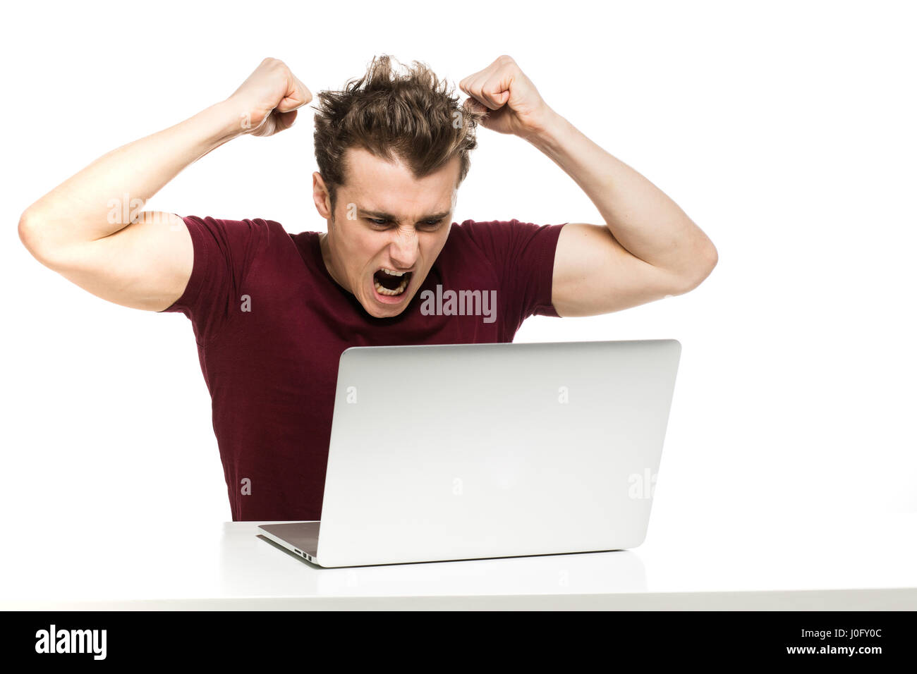 young man in red shirt sitting at table with laptop in front being very angry, isolated on white Stock Photo