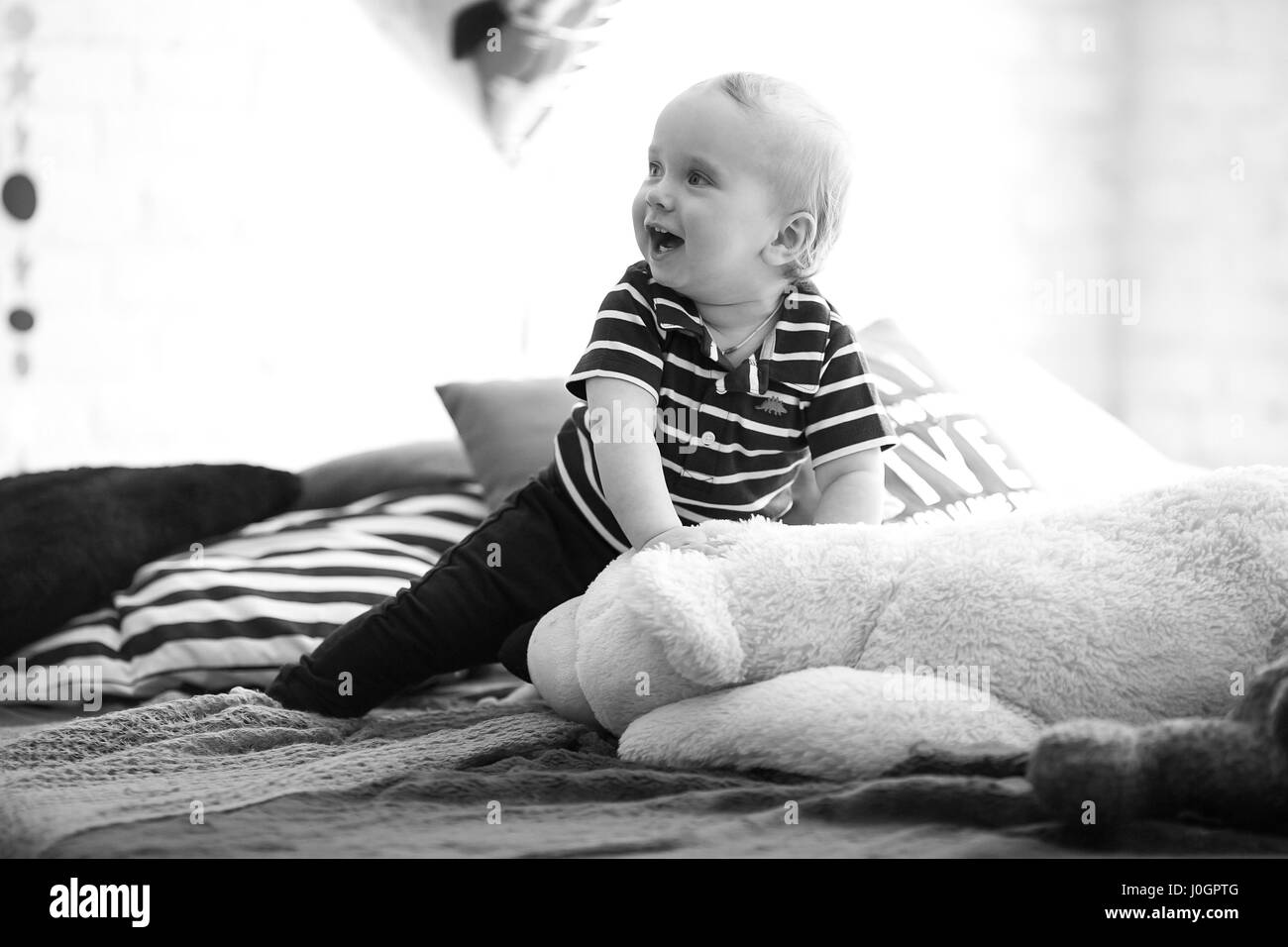 Small baby sits on bed next to teddy bear and laughs. Black and white image. Stock Photo