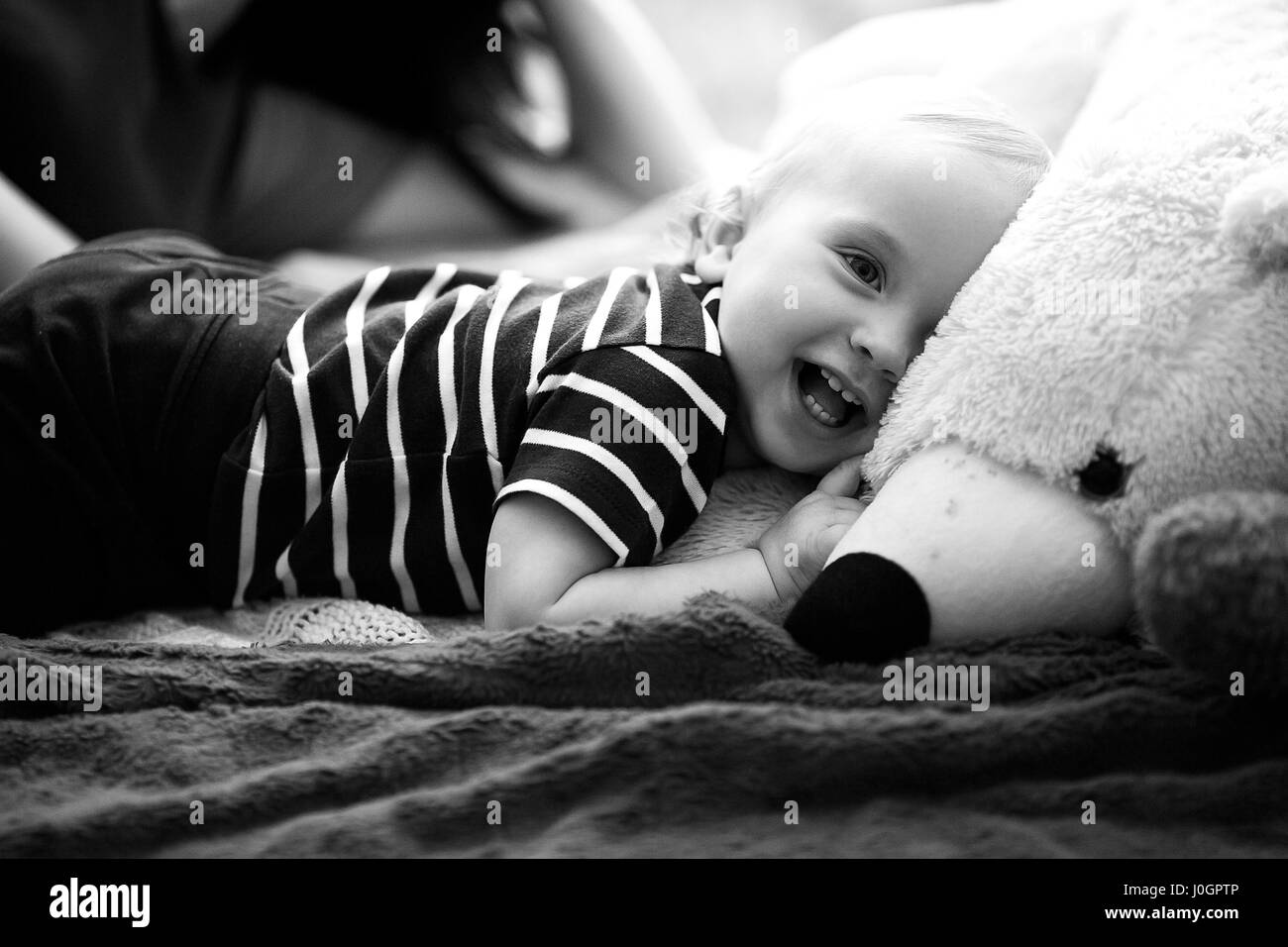 Small baby lies on bed next to teddy bear and laughs. Black and white image. Stock Photo