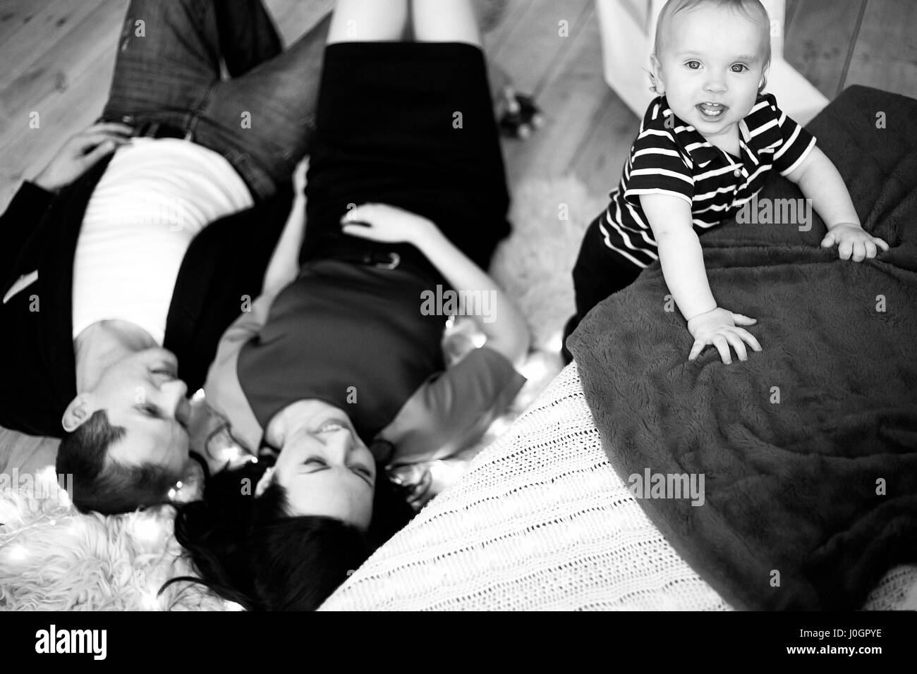 Young parents lie on carpet, baby stands nearby and smiles. Black and white image. Stock Photo