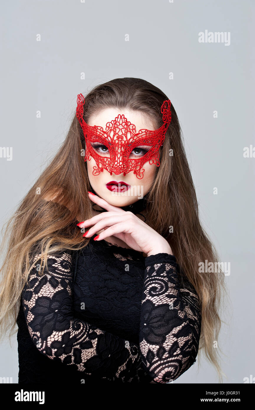 Young woman with fair hair, wearing a red lace mask and a black dress with lace sleeves Stock Photo