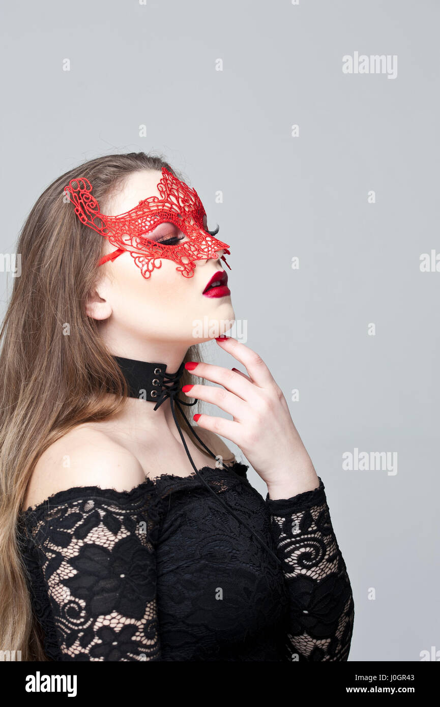 Young woman with fair hair, wearing a red lace mask and a black dress with lace sleeves Stock Photo
