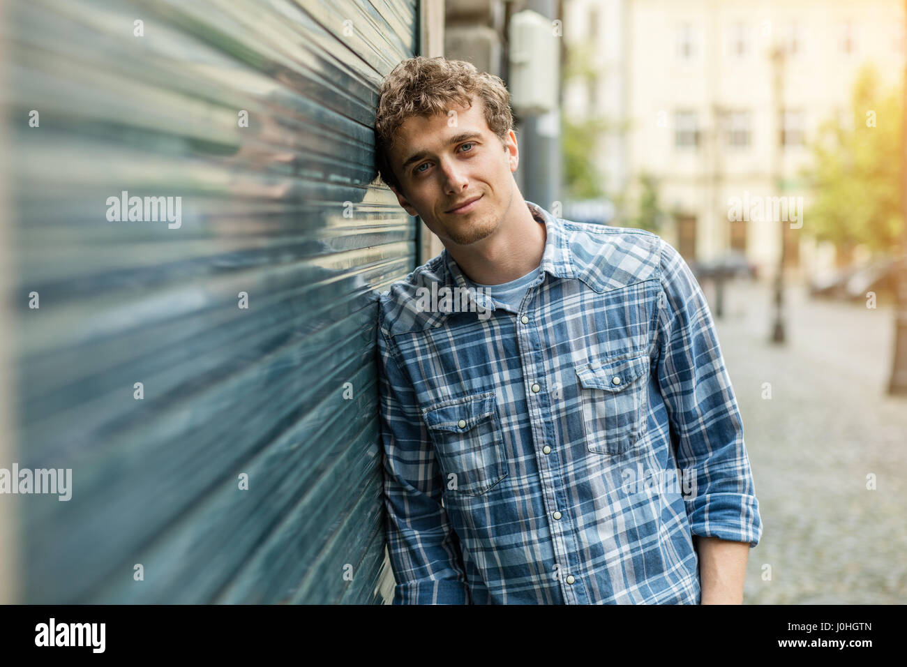 young blond and culry man in checkered shirt standin outside next to a wall Stock Photo