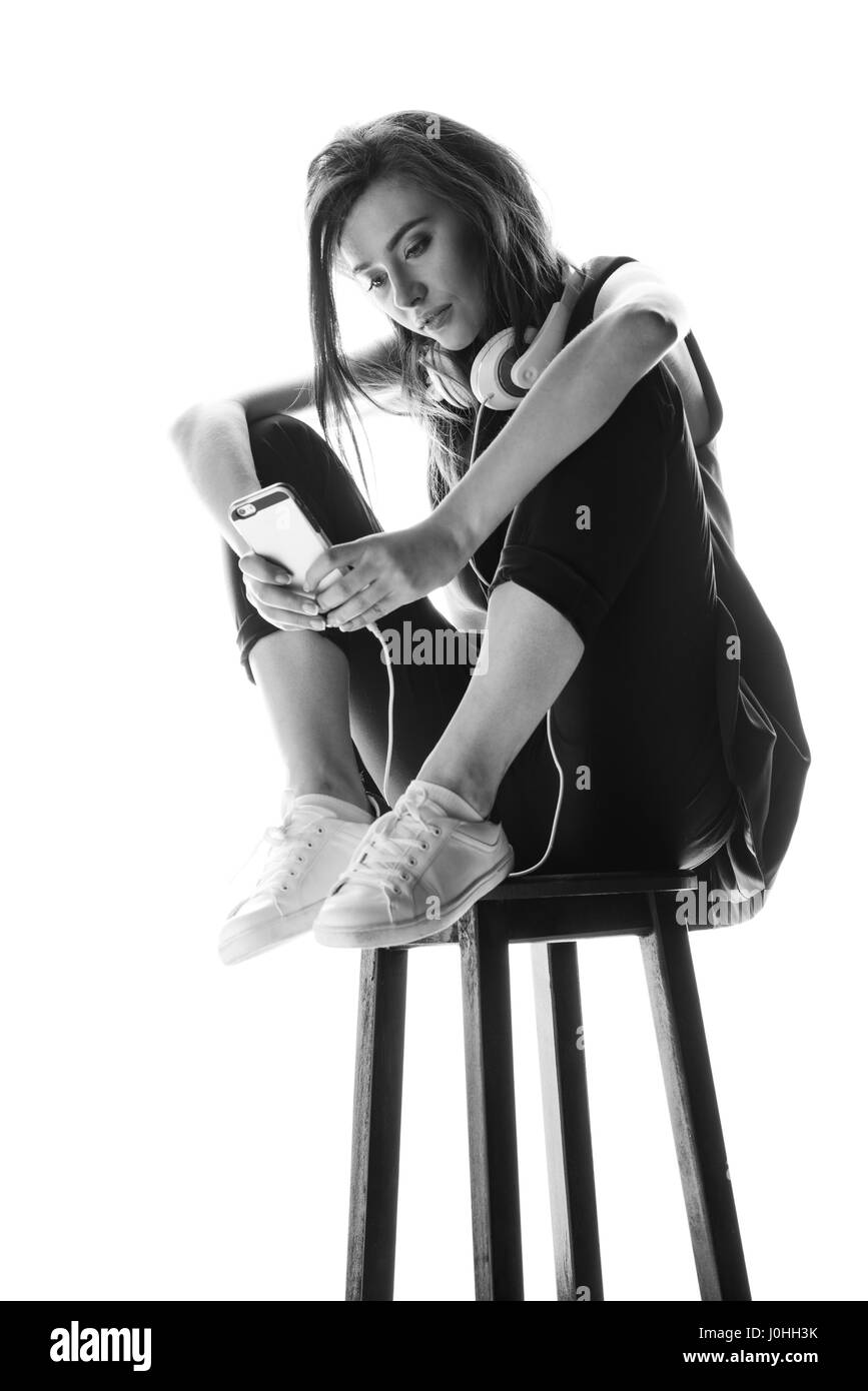 Black and white photo of young woman with headphones sitting on stool and using smartphone Stock Photo