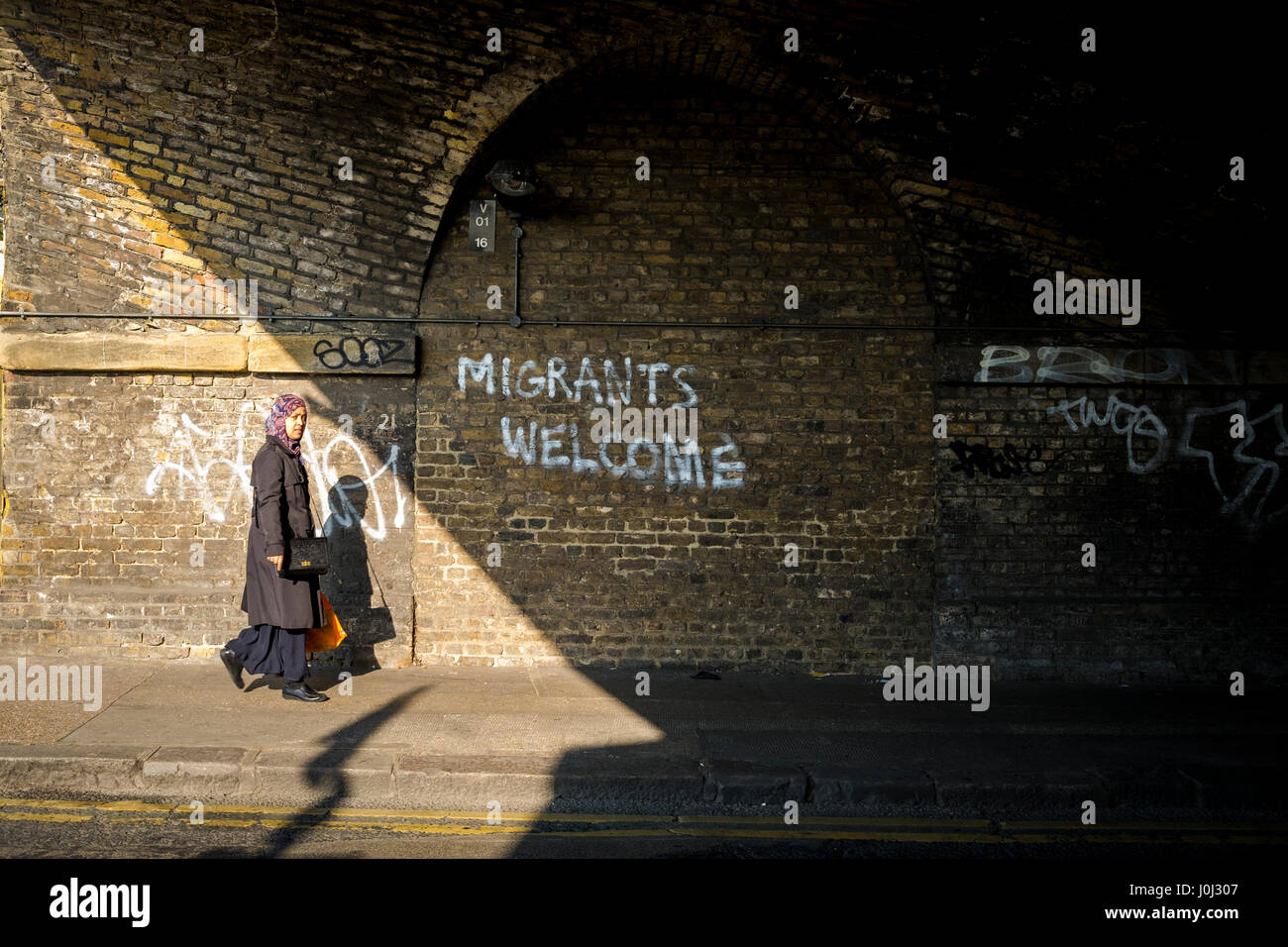 Asian woman walks past graffiti in London's Bethnal Green welcoming migrants. Stock Photo