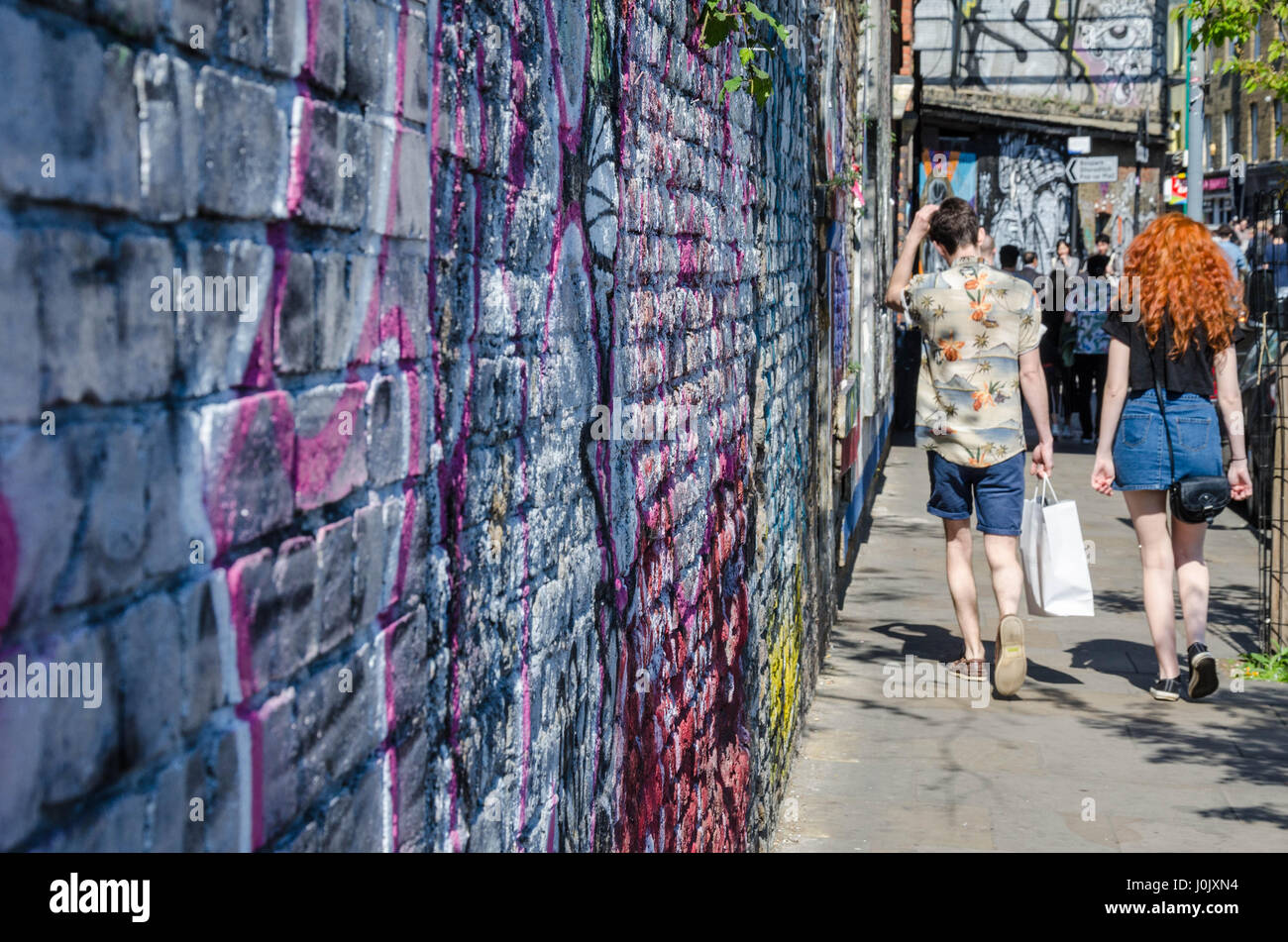 A couple of pedestrians walk down a footpath alongside a brick wall on Brick Lane which is covered in street are and graffiti. Stock Photo