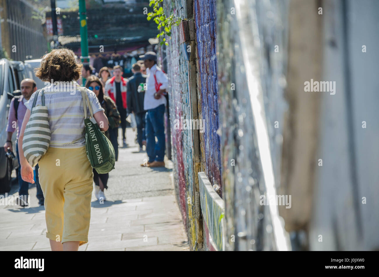 Pedestrians walk down a footpath alongside a brick wall on Brick Lane which is covered in street are and graffiti. Stock Photo