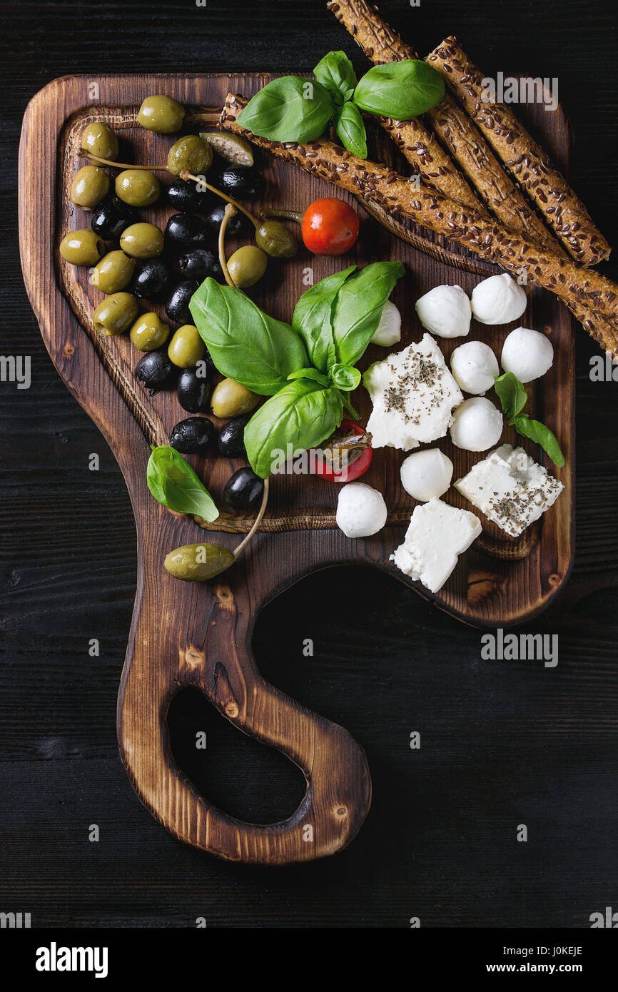 Mediterranean appetizer antipasti board with green black olives, feta cheese, mozzarella, capers, pepper, basil with grissini bread sticks over black  Stock Photo