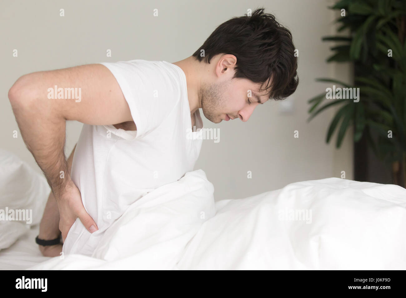 Young man sitting on bed at home touching aching back Stock Photo