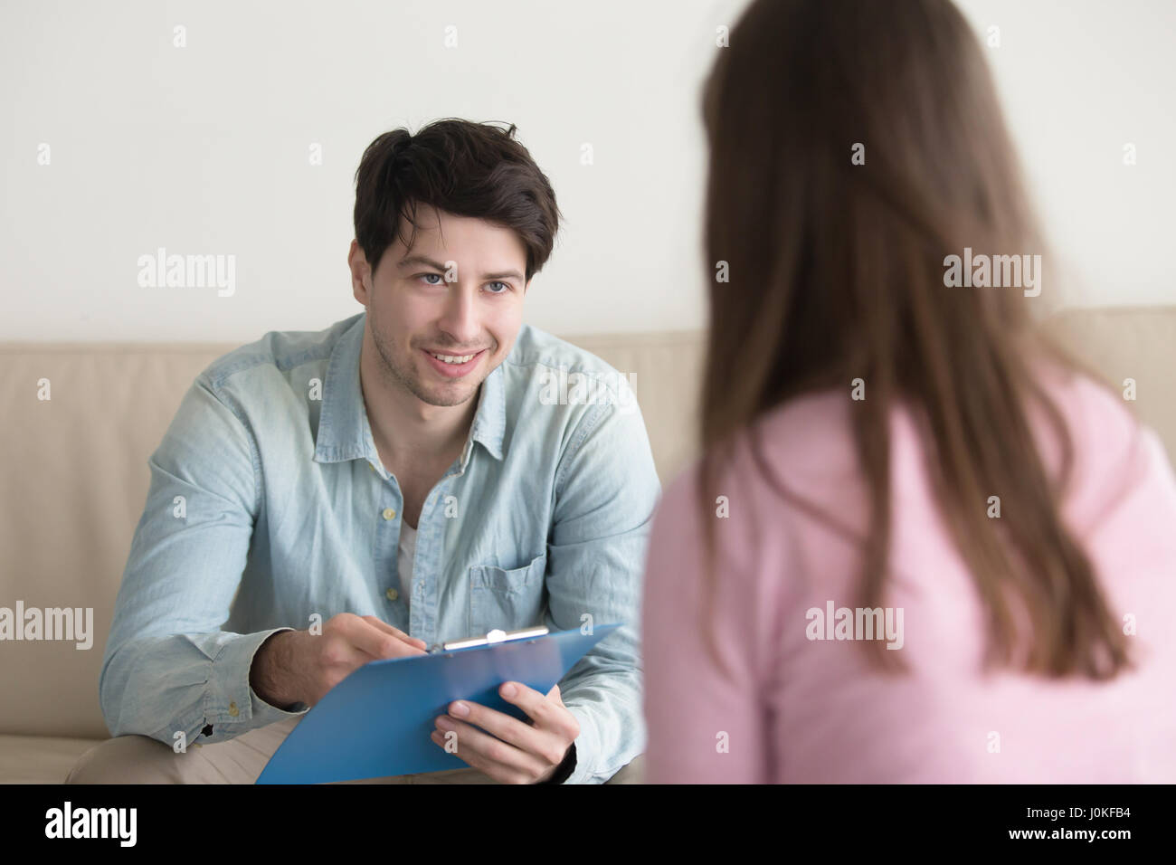 Cheerful man holding clipboard sitting opposite woman, asking or Stock Photo