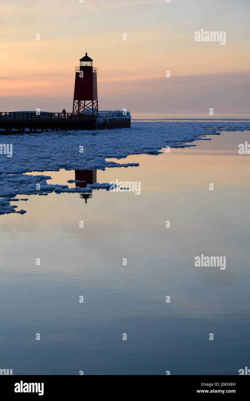 A lighthouse reflects in the calm snow and ice filled water of Lake Michigan at sunset in the winter. Stock Photo