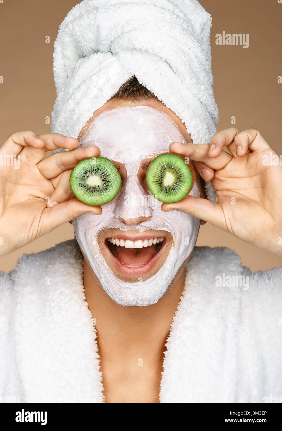 Funny young man with facial mask and pieces of kiwis on eyes. Photo of well groomed man receiving spa treatments. Beauty & Skin care concept Stock Photo