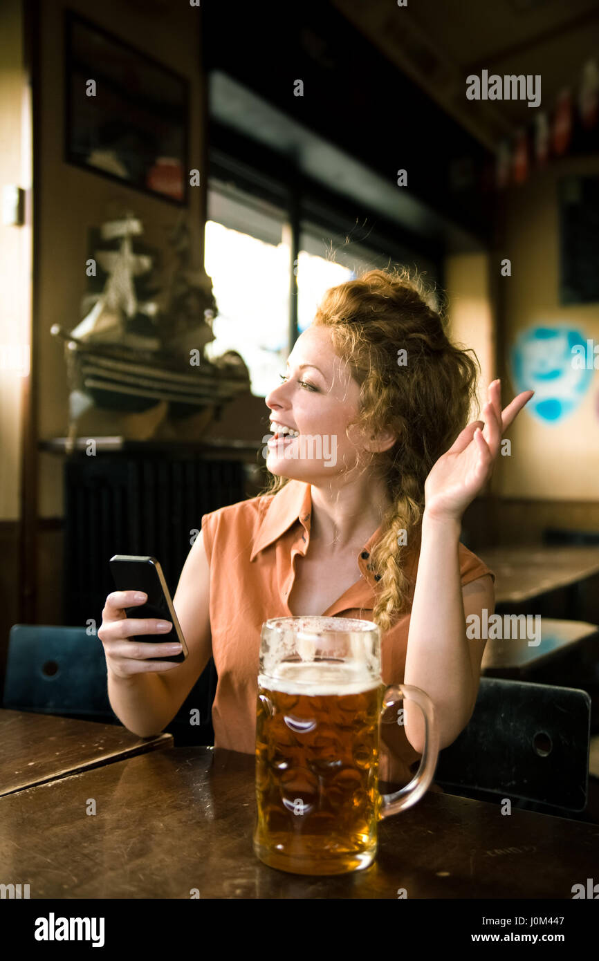 Blonde young woman waving at someone outside the frame. She is holing a callphone and has a glass of beer in front of her on the table Stock Photo