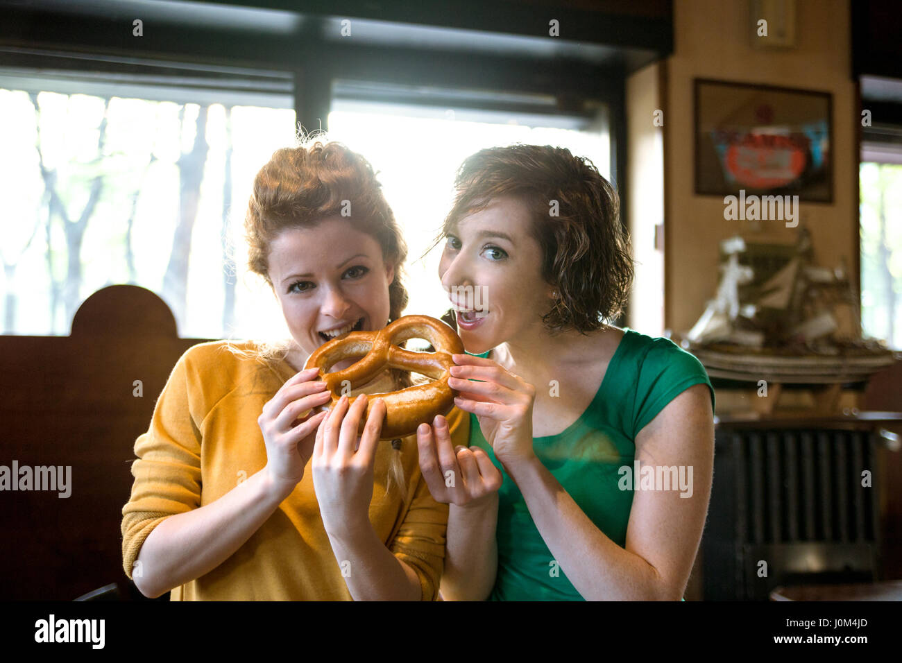 Two girlfriends eating pretzel, looking at camera indoor setting. Both are smiling and enjoying their time together Stock Photo