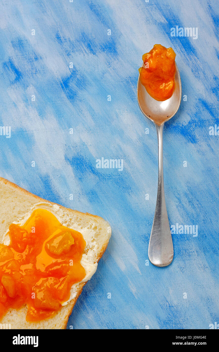 Toast slice with butter and jam and silver spoon with jam blob over blue painted textile background. Overhead view. Stock Photo