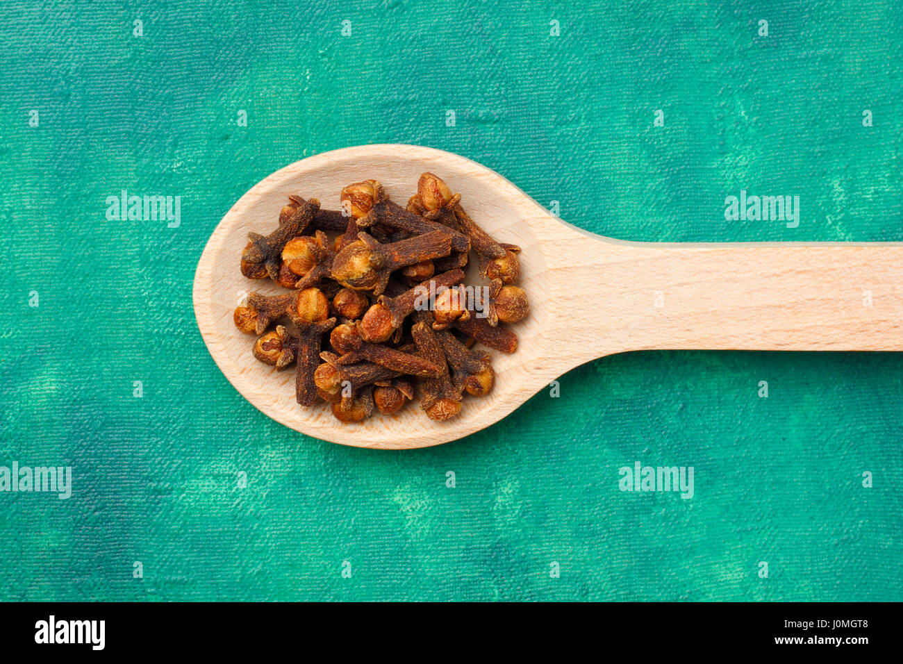 Cloves (Syzygium aromaticum) dried flower buds on wooden spoon over emerald painted textile background Stock Photo