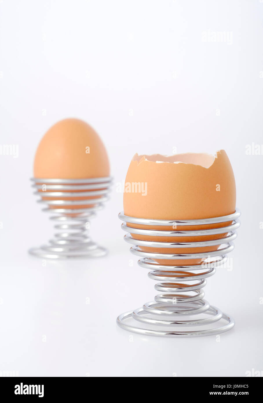 Two eggs in wire eggcups on bright background. Focus on front egg with partially removed eggshell. Copy space at top of photo. Stock Photo