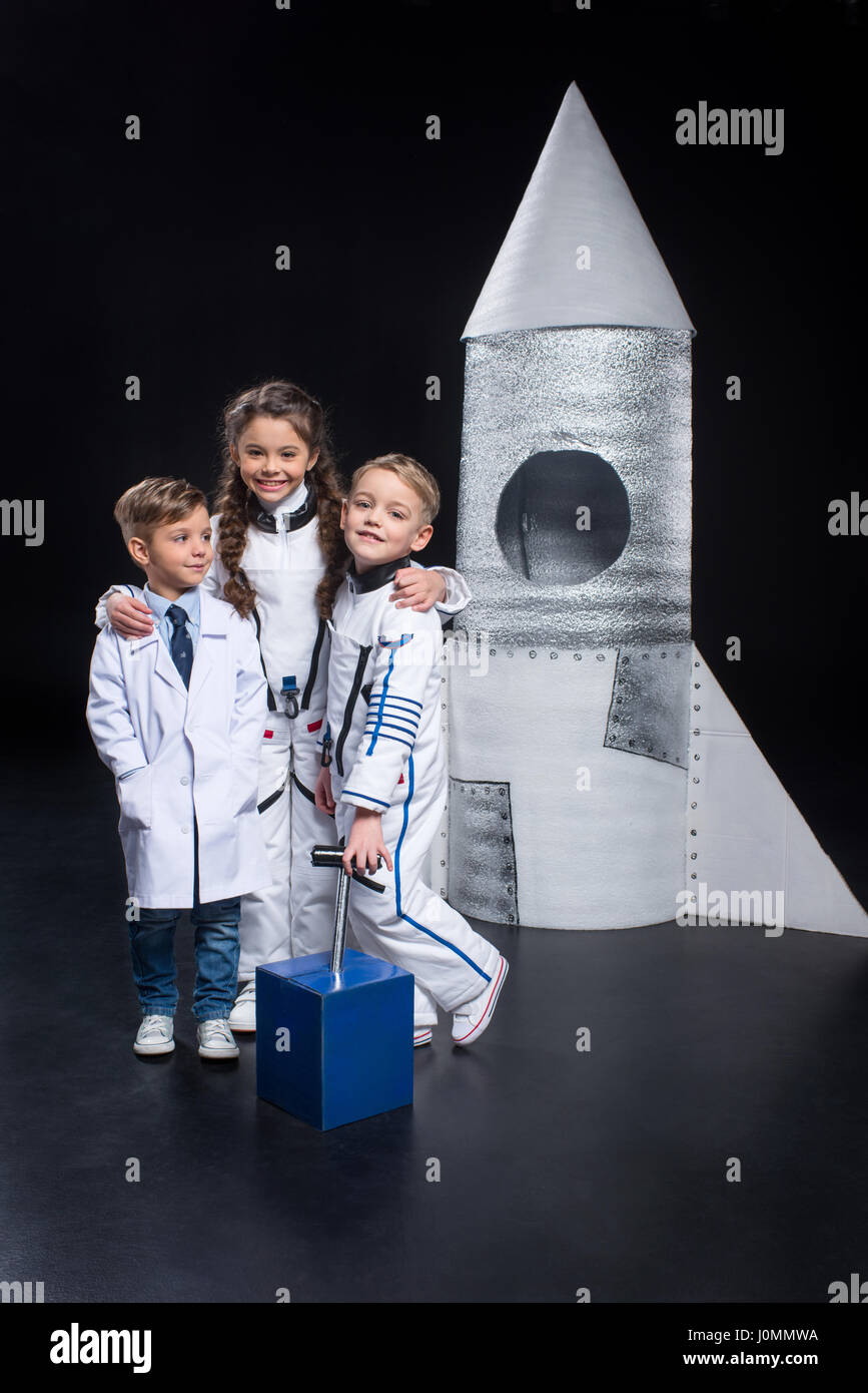 Three smiling little kids hugging and playing astronauts with rocket on black Stock Photo