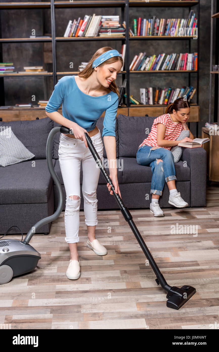 Smiling young woman cleaning floor with vacuum cleaner while female ...