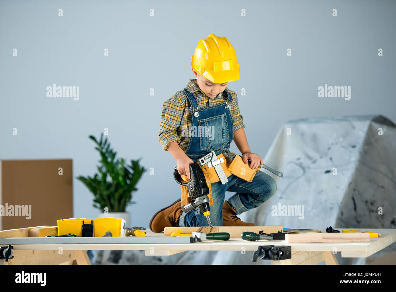 Cute little boy in helmet and tool belt drilling wooden plank with toy