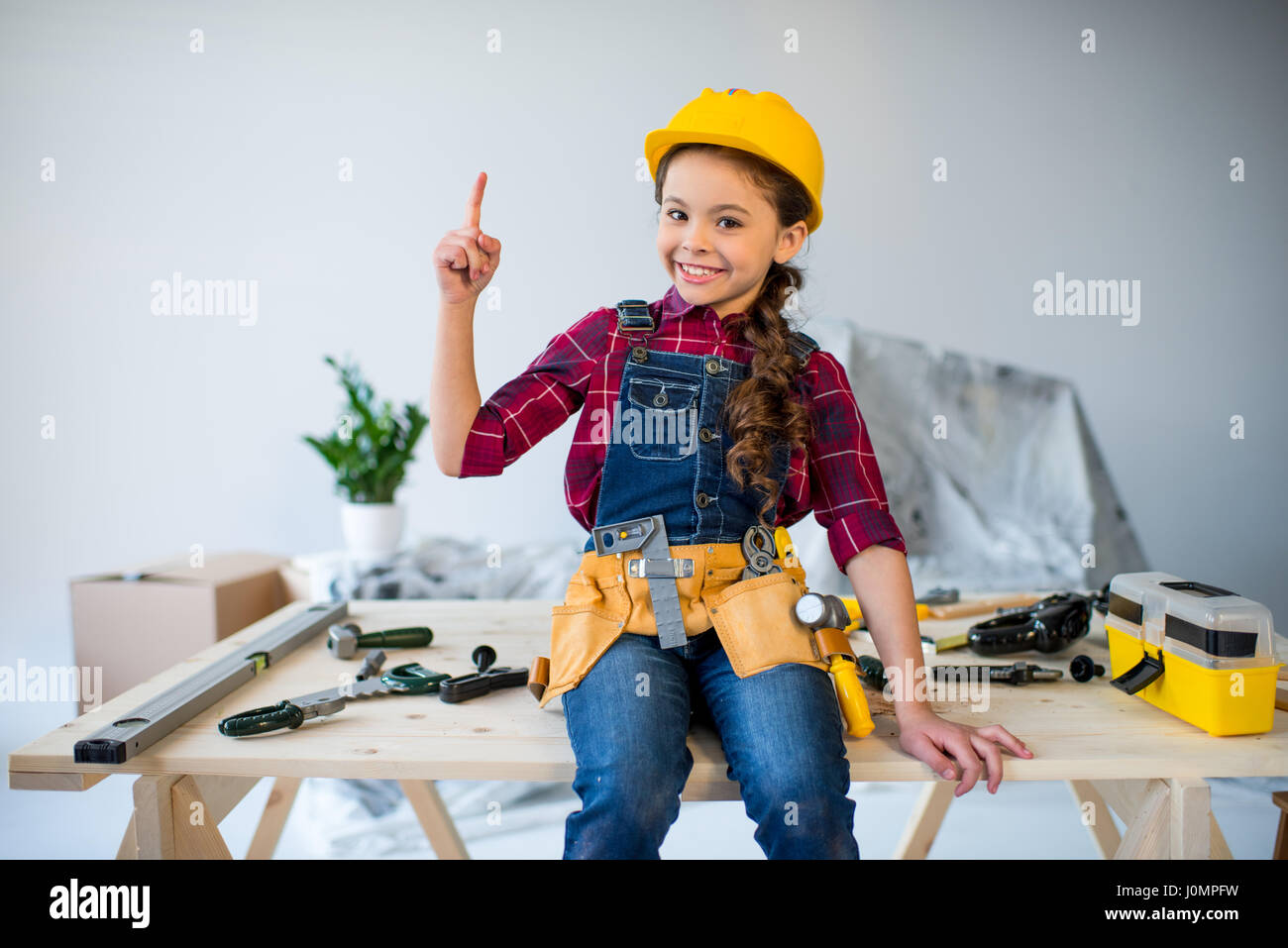 Little girl in tool belt and hard hat pointing up and smiling at camera