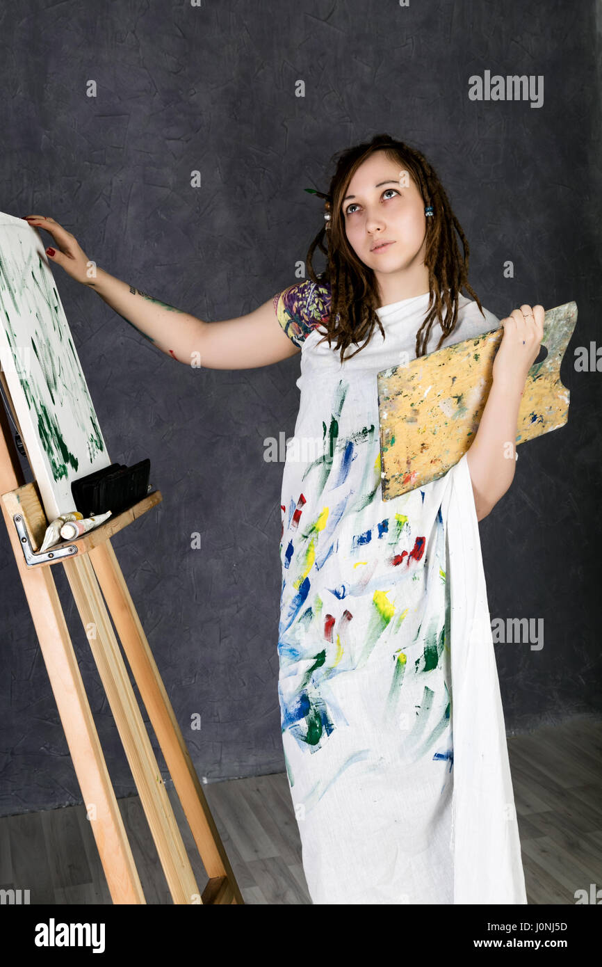 female paint artist posing next to a easel and paints on an canvas ...