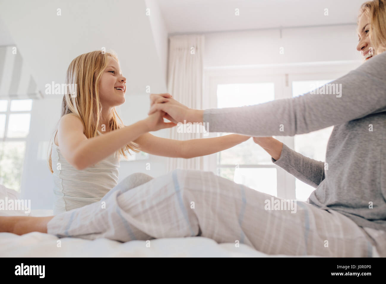 Indoor shot of little girl playing with her mother on bed. Mother and daughter sitting in bedroom holding hands. Stock Photo