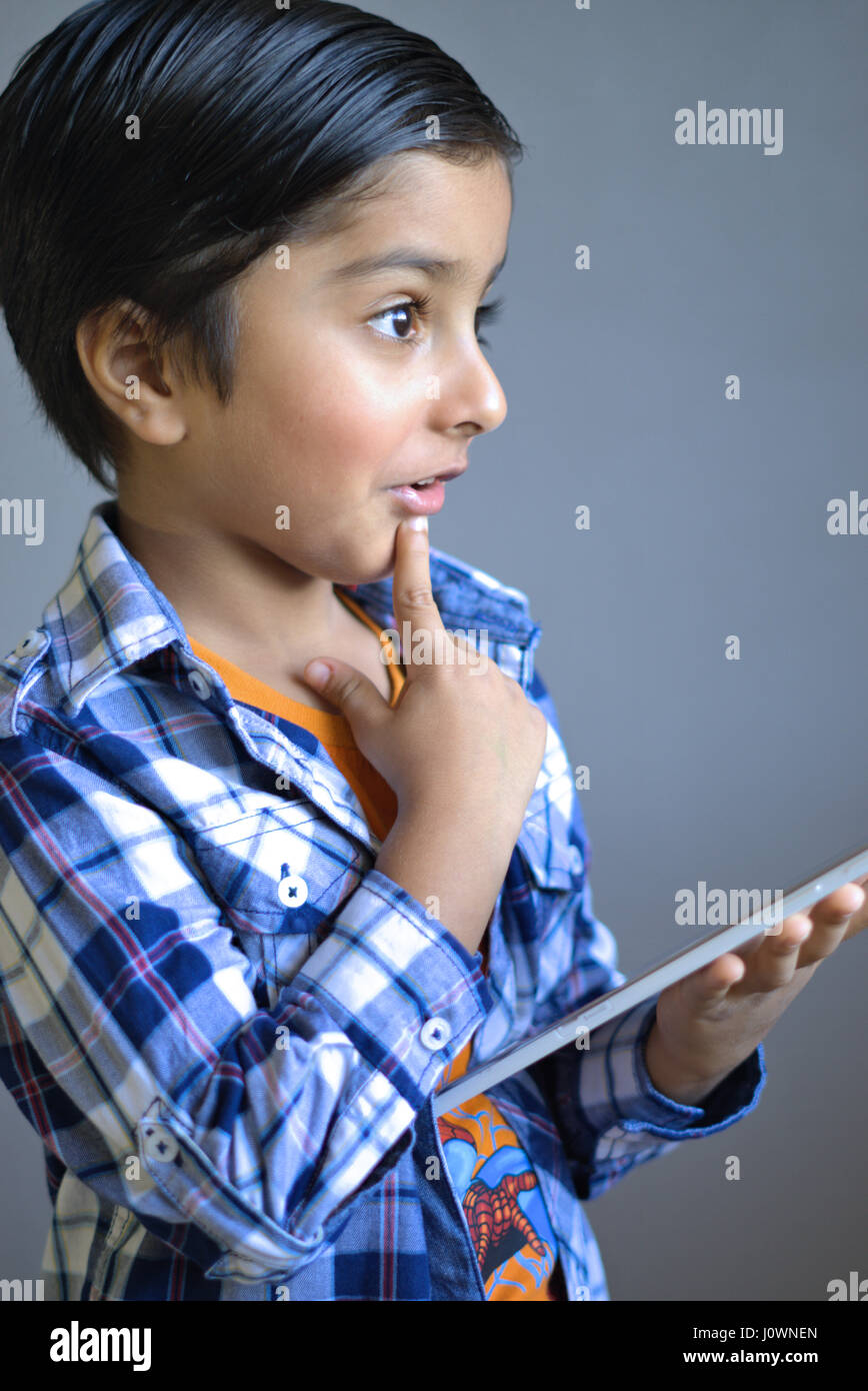 Kid with tablet computer. Kid thinking holding tablet. Kid holding a tablet device with his finger on his chin as if thinking and wondering. Stock Photo