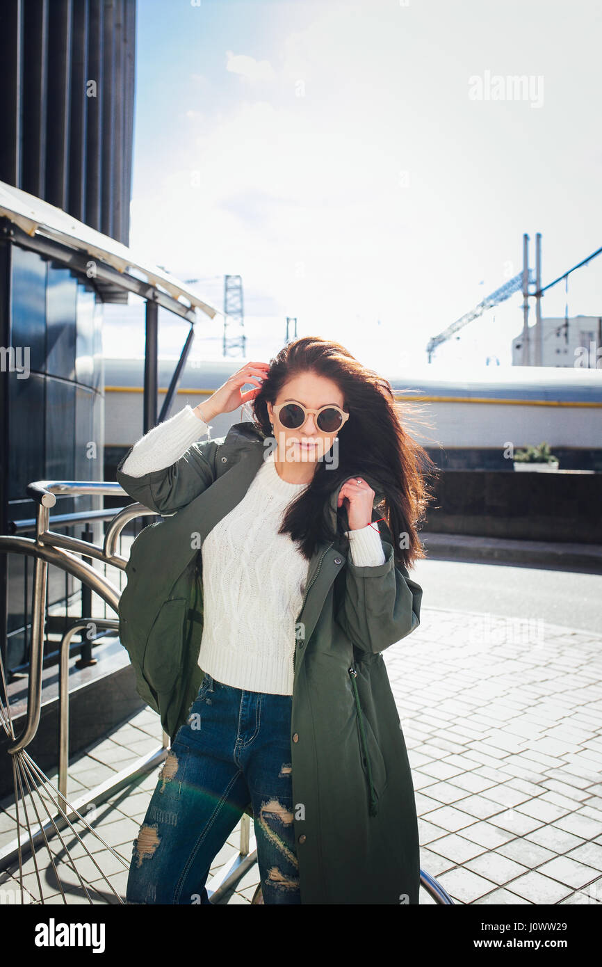 fashion closeup portrait of nice pretty young hipster woman posing in sunglasses Outdoor .Brunette happy girl in green raincoat and pink sneakers walks the street of the city. Stock Photo