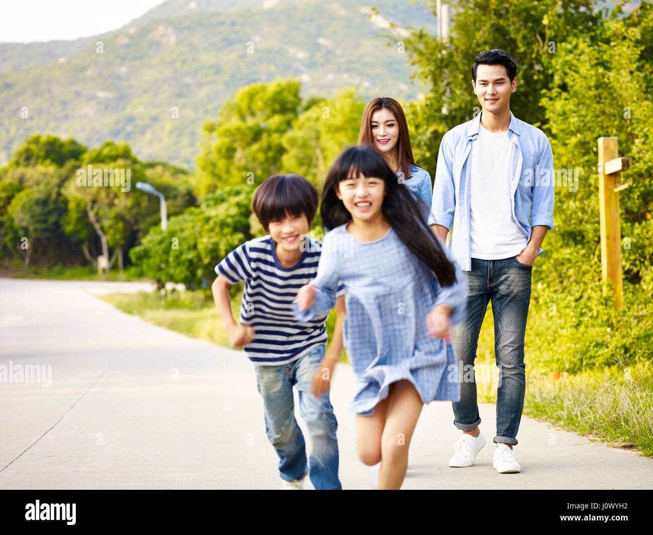 two asian children running in park while their parents watching affectionately. Stock Photo