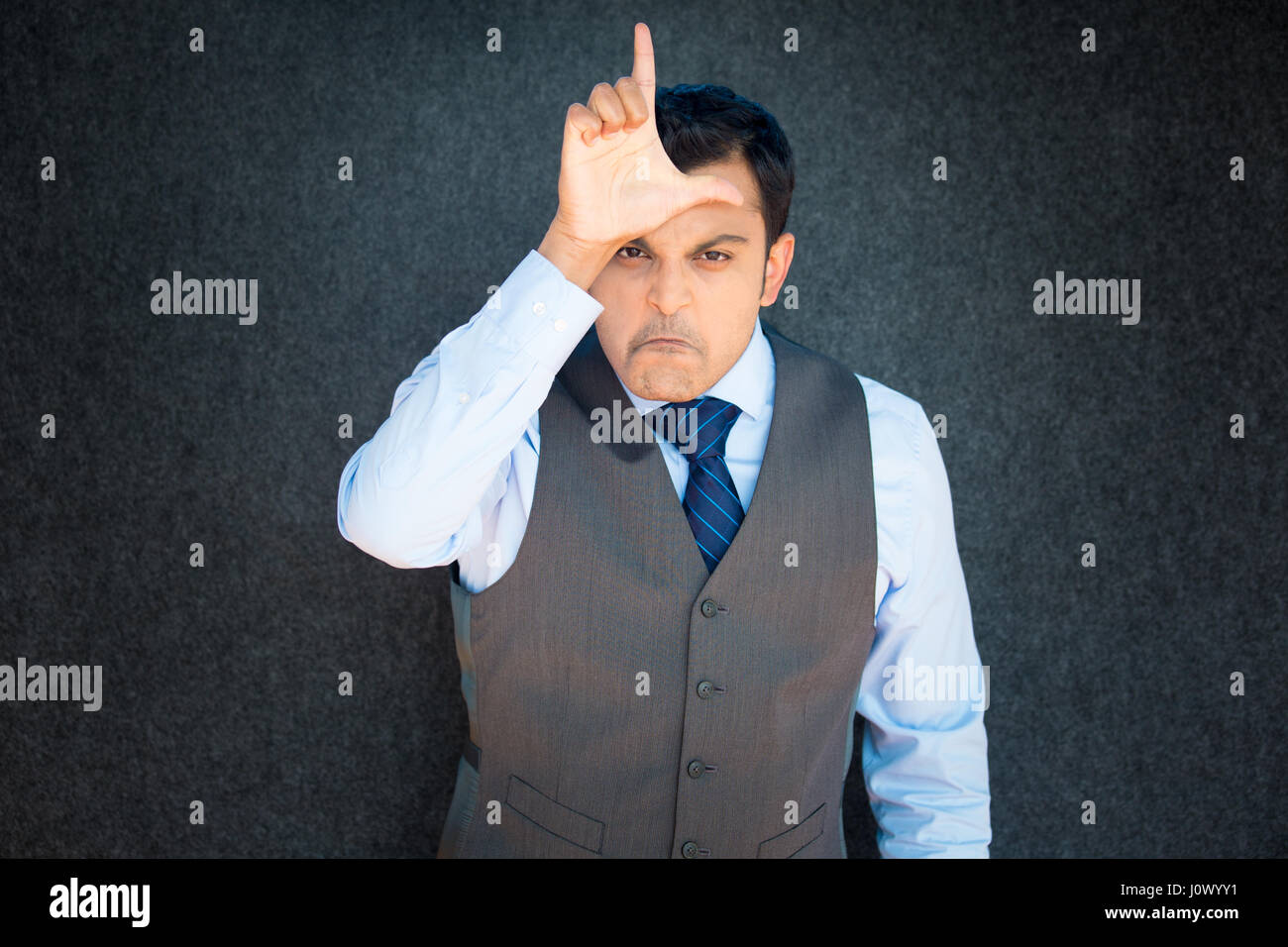 Closeup portrait, funny young man in vest and tie displaying loser sign on forehead, looking at you with disgust, isolated gray black background. Nega Stock Photo