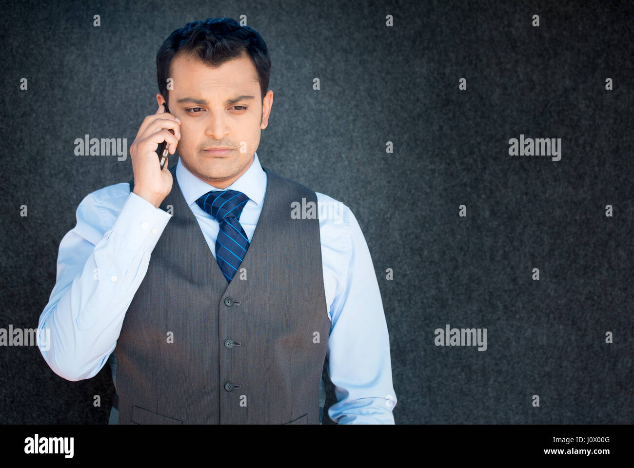 Closeup portrait, unhappy worried young man in vest and tie talking on phone, isolated gray black background. Negative human emotions, facial expressi Stock Photo