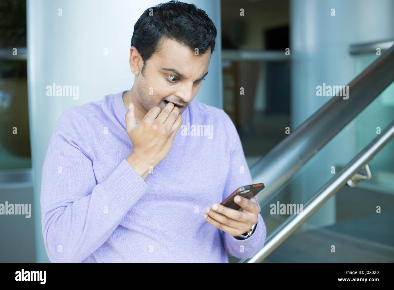 Closeup portrait, funny young man, shocked surprised, biting fingernails, by what he sees on his cell phone, isolated indoors office background. Negat Stock Photo