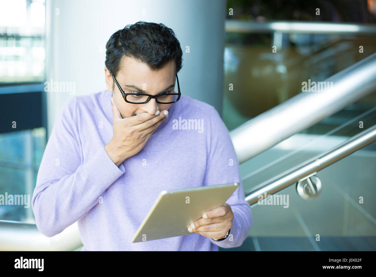 Closeup portrait, young happy man in black eyeglasses and purple sweater, astonished, hand on mouth concealed laugh, by what he sees on tablet, isolat Stock Photo