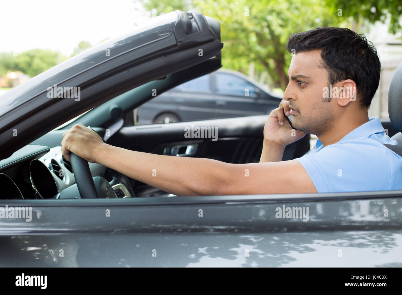 Closeup portrait, sad handsome young man in blue polo shirt sitting, driving in gray black sports car on cellphone with bad news, isolated outdoors ba Stock Photo