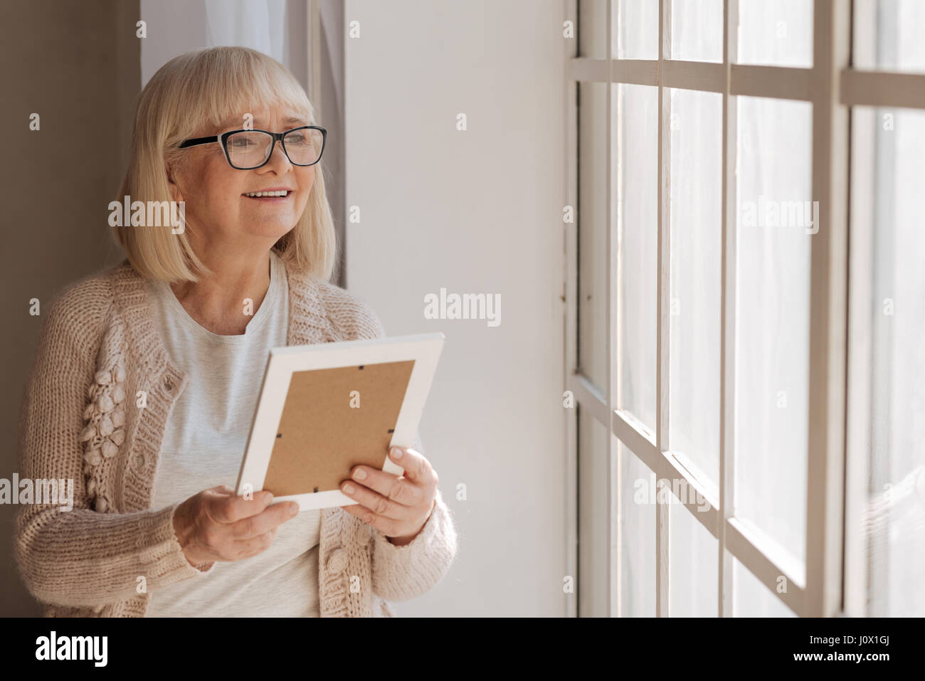 Positive mood. Delighted nice aged woman looking into the window and smiling while holding a photograph Stock Photo