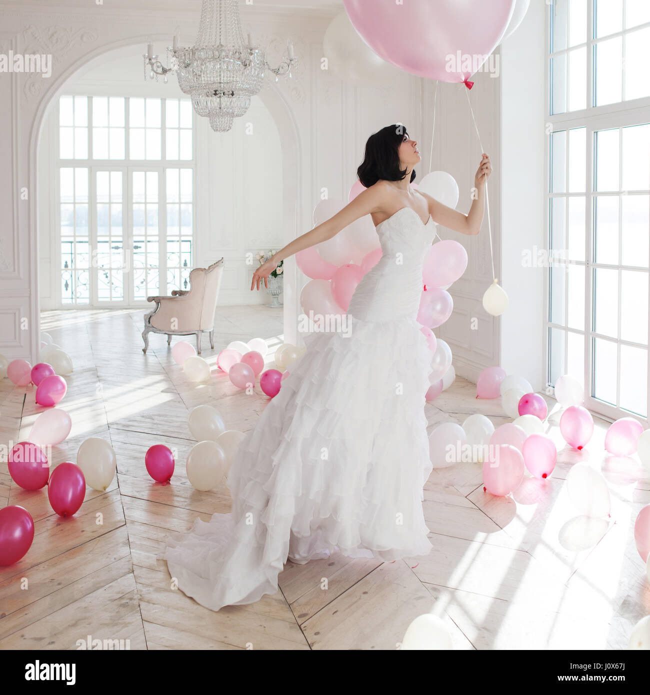 Young woman in wedding dress in luxury interior flies on pink and white balloons. Stock Photo