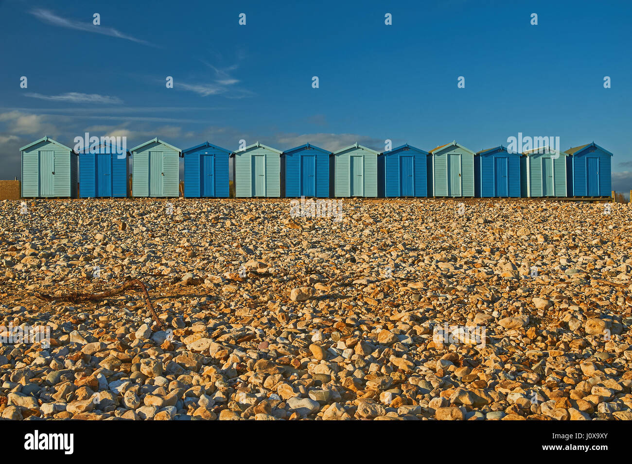 Charmouth, Dorset and a line of beach huts in alternating light and dark blue colours at the back of the shingle beach on a summer evening. Stock Photo