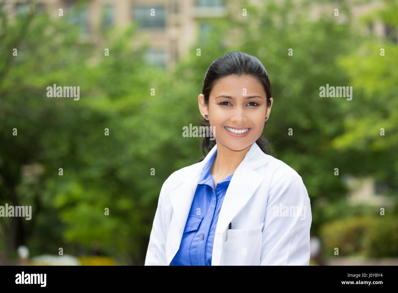 Closeup headshot portrait of friendly, cheerful, smiling confident female, healthcare professional with lab coat. isolated outdoors outside green tree Stock Photo