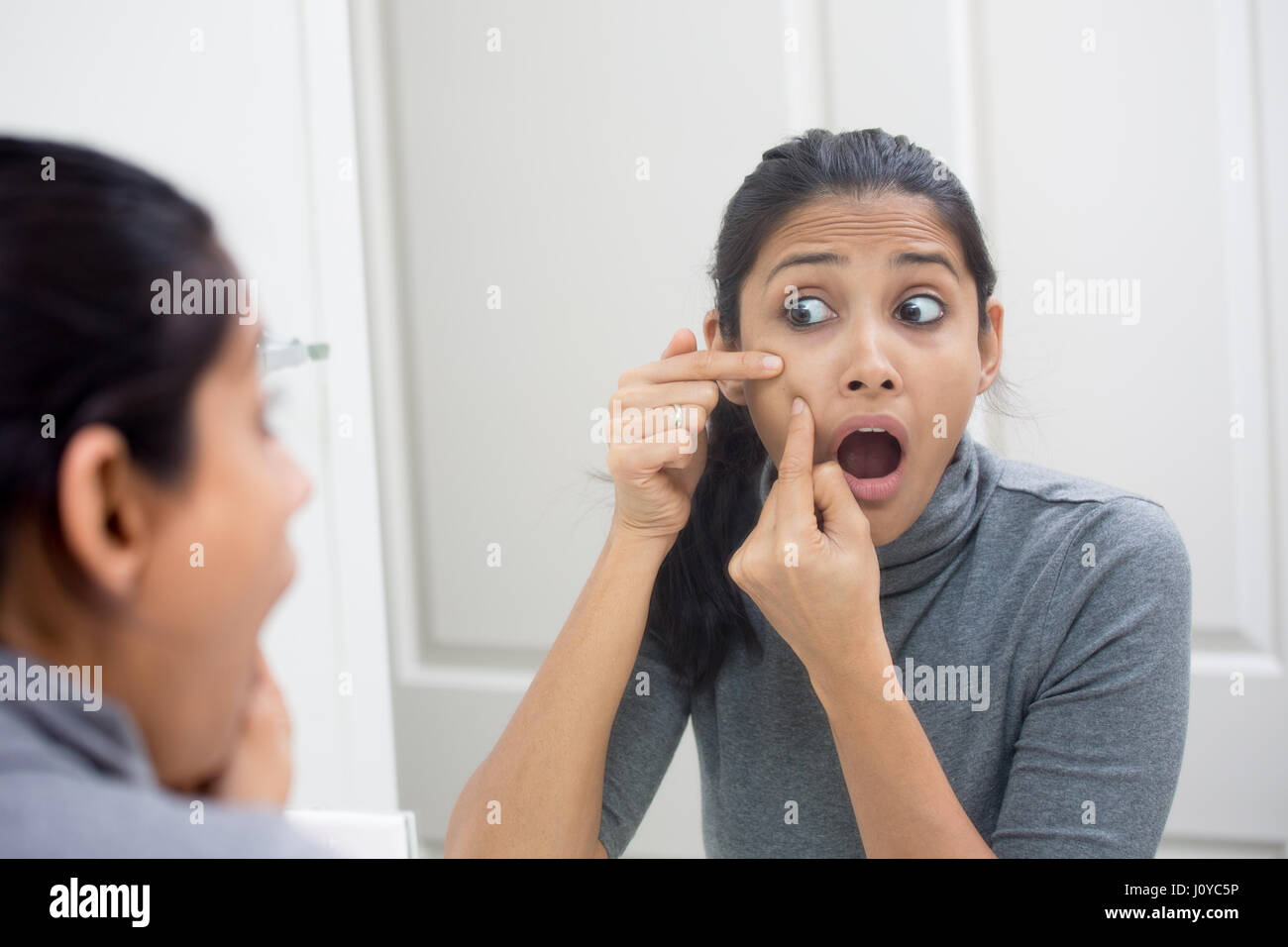 Closeup portrait of young frustrated woman surprised stunned to see zit on her face, gray turtleneck, isolated mirror reflection background. Facial  f Stock Photo